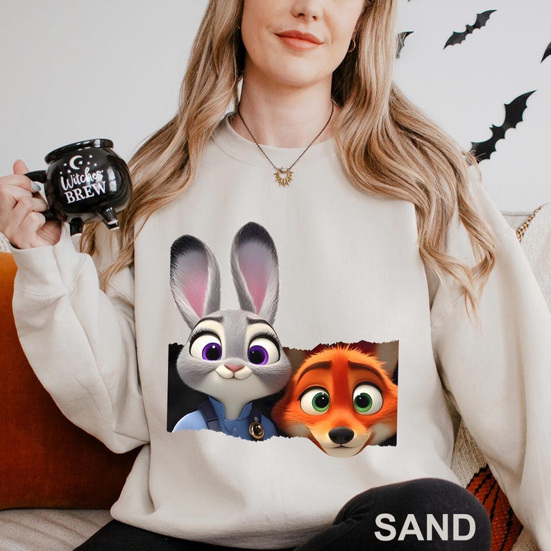 Może przedstawiać: Beżowa bluza z nadrukiem Judy Hopps i Nicka Wilde z filmu Zwierzogr&oacute;d. Projekt przedstawia postacie wyglądające przez efekt podartego papieru. Osoba na zdjęciu trzyma czarny kubek z napisem "Witches Brew".
