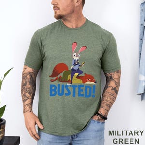 May include: Military green t-shirt with a cartoon image of a rabbit police officer and a fox, and the word "BUSTED!" in blue. The shirt is made of a soft, comfortable material.