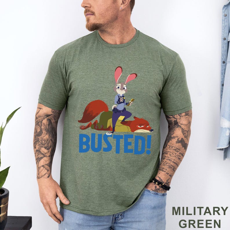 May include: Military green t-shirt with a cartoon image of a rabbit police officer and a fox, and the word "BUSTED!" in blue. The shirt is made of a soft, comfortable material.