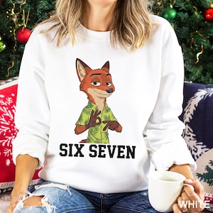 May include: White sweatshirt featuring a cartoon fox character wearing a green shirt. The text "SIX SEVEN" is printed in black block letters below the fox. The sweatshirt is a casual style.