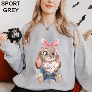 May include: Sport grey sweatshirt with a cartoon bunny holding a heart that says "Love". The bunny has a pink bow and wears a police uniform. The sweatshirt has the words "SPORT GREY" printed at the top.