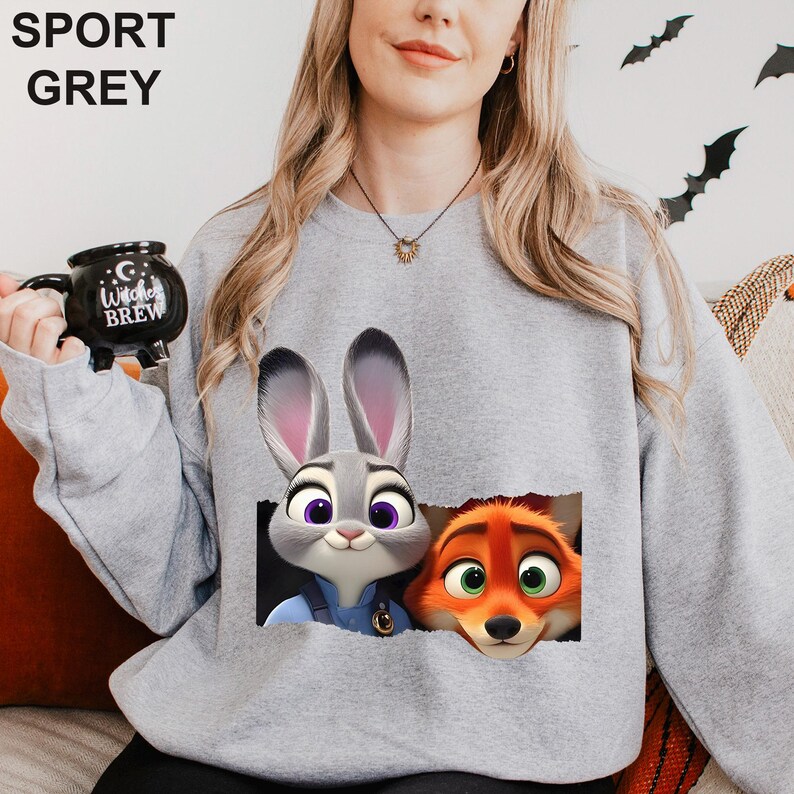 Może przedstawiać: Szara bluza sportowa z grafiką Judy Hopps i Nicka Wilde z Zootopii. Bluza ma okrągły dekolt i długie rękawy. Tekst "SPORT GREY" jest wydrukowany u g&oacute;ry. Czarny kubek z napisem "Witches Brew" jest trzymany w lewej ręce.