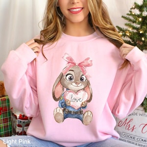 May include: Pale pink sweatshirt with a watercolour illustration of a bunny holding a heart-shaped sign that says "Love". The bunny has a pink bow and is wearing a blue uniform. The text "Light Pink" is at the bottom left.