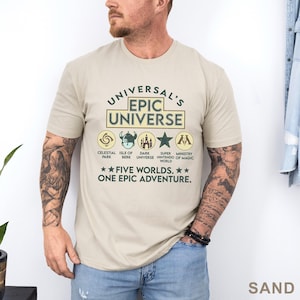 May include: Sand-colored t-shirt with the text "UNIVERSAL'S EPIC UNIVERSE" and various themed icons. Below the text are the words "FIVE WORLDS. ONE EPIC ADVENTURE."