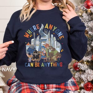 May include: Navy blue sweatshirt featuring a colorful graphic of a city skyline and various animal characters. The text "WHERE ANYONE CAN BE ANYTHING" is displayed in a rainbow font. The word "NAVY" is printed on the bottom left.