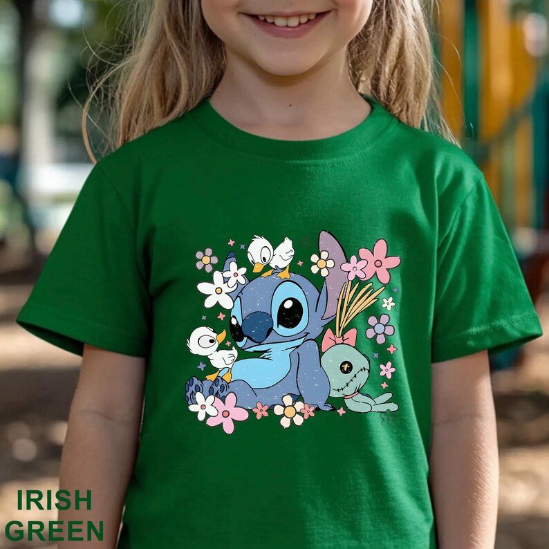 K&ouml;nnte beinhalten: Gr&uuml;nes T-Shirt mit einer Grafik eines blauen Zeichentrickcharakters, umgeben von Blumen und kleinen wei&szlig;en V&ouml;geln. Der Text "IRISH GREEN" befindet sich unten links auf dem Shirt.