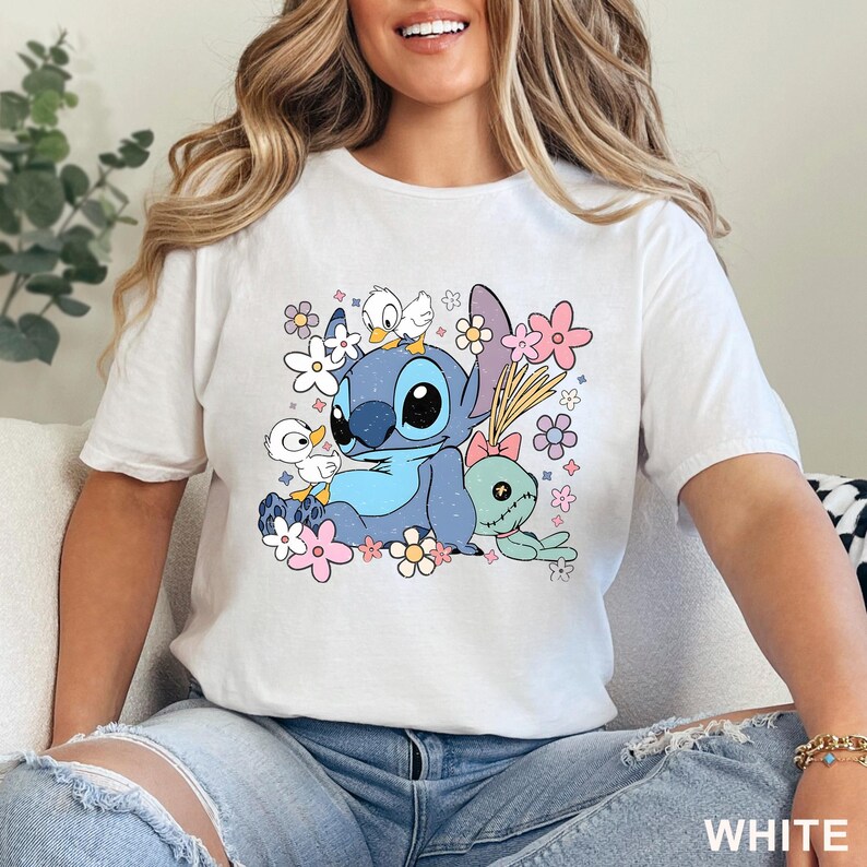 K&ouml;nnte beinhalten: Wei&szlig;es T-Shirt mit einem farbenfrohen Aufdruck von Stitch aus Lilo & Stitch, umgeben von Blumen und anderen Charakteren. Das Design umfasst Blau-, Rosa- und Gelbt&ouml;ne. Das Wort "WHITE" ist unten rechts aufgedruckt.