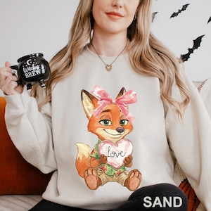 May include: Sand-coloured sweatshirt with a cartoon fox holding a heart that says "Love." The fox wears a pink bow, a Hawaiian shirt, and khaki shorts. A black mug with "Witches Brew" is held in the background.