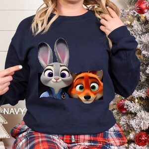 Może przedstawiać: Granatowa bluza z nadrukiem Judy Hopps, kr&oacute;lika, i Nicka Wilde, lisa, z filmu Zwierzogr&oacute;d. Postacie są przedstawione w kieszeni. Słowo "NAVY" jest wydrukowane po lewej stronie bluzy.