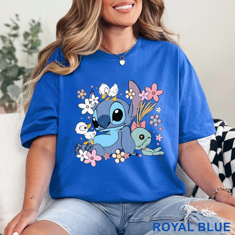 K&ouml;nnte beinhalten: K&ouml;nigsblaues T-Shirt mit einem Cartoon-Stitch-Design, umgeben von Blumen und kleinen Enten. Das Design zeigt Stitch, eine gr&uuml;ne Puppe und pastellfarbene Blumen. Das Shirt hat eine leuchtende k&ouml;nigsblaue Farbe.