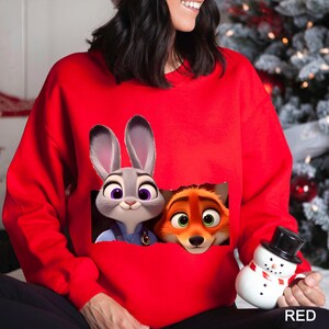Może przedstawiać: Czerwona bluza z nadrukiem Judy Hopps i Nicka Wilde z filmu Zwierzogr&oacute;d. Postacie z kresk&oacute;wek wyglądają z kieszeni. Słowo "RED" jest wydrukowane na dole po prawej stronie.