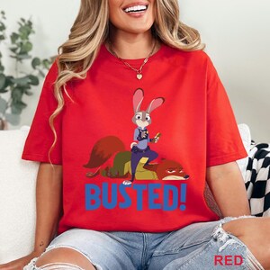 May include: Red t-shirt with a cartoon illustration of Judy Hopps and Nick Wilde from Zootopia. The text "BUSTED!" is printed in blue below the characters. The word "RED" is printed in the bottom right corner of the t-shirt.
