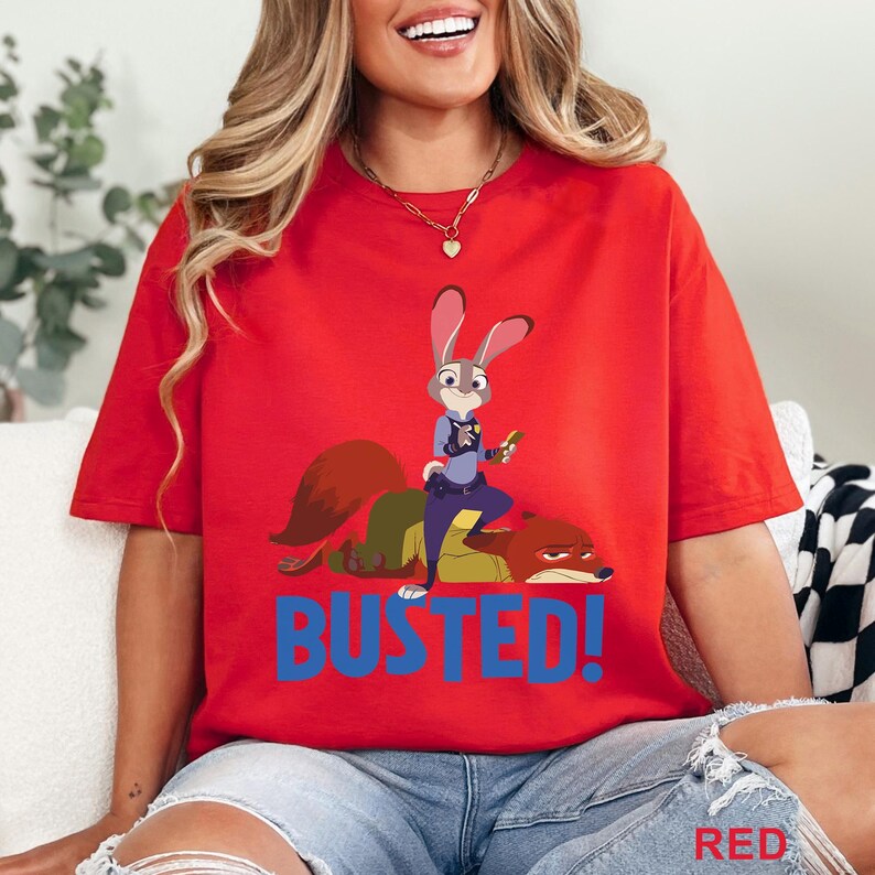 May include: Red t-shirt with a cartoon illustration of Judy Hopps and Nick Wilde from Zootopia. The text "BUSTED!" is printed in blue below the characters. The word "RED" is printed in the bottom right corner of the t-shirt.