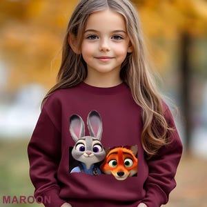 Może przedstawiać: Bordowa bluza z postaciami z kresk&oacute;wki Judy Hopps i Nickiem Wilde z Zwierzogrodu. Bluza ma okrągły dekolt i długie rękawy. Słowo "MAROON" jest wydrukowane na dole obrazu.