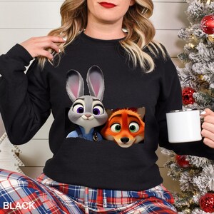 Może przedstawiać: Czarna bluza z grafiką Judy Hopps i Nicka Wilde z filmu Zwierzogr&oacute;d. Postacie znajdują się w kieszeni. Słowo "BLACK" jest wydrukowane w lewym dolnym rogu obrazu.