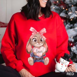 May include: A red sweatshirt with a cartoon bunny holding a heart-shaped sign with the word "Love" on it. The bunny has a pink bow and is wearing a blue uniform. The word "RED" is printed in white at the bottom right.