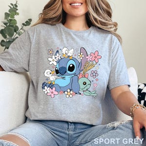 K&ouml;nnte beinhalten: Graues T-Shirt mit einem farbenfrohen Aufdruck von Stitch, umgeben von Blumen und kleinen Entenk&uuml;ken. Der Text "SPORT GREY" ist unten auf dem Shirt aufgedruckt. Das Design ist in Blau-, Rosa- und Gr&uuml;nt&ouml;nen gehalten.