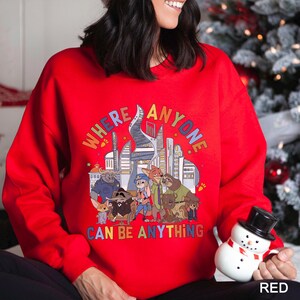 May include: A red sweatshirt with a colorful graphic featuring cartoon characters and the text "WHERE ANYONE CAN BE ANYTHING". The design includes a cityscape and various animal figures.
