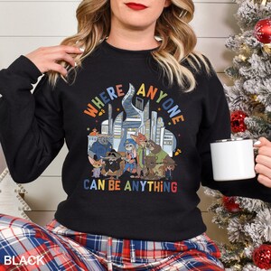 May include: Black crewneck sweatshirt featuring a colorful graphic of cartoon characters and the text "WHERE ANYONE CAN BE ANYTHING". The design includes a cityscape and various animal characters. The sweatshirt is a comfortable, casual style.
