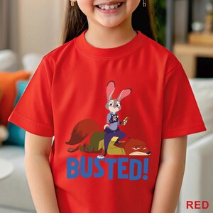 May include: A red t-shirt with a cartoon image of Judy Hopps and Nick Wilde from Zootropolis. The word "BUSTED!" is printed in blue below the characters. The word "RED" is printed in the bottom right corner of the t-shirt.