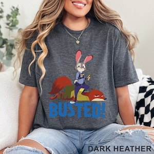 May include: Dark heather grey t-shirt with a cartoon image of Judy Hopps, a bunny police officer, standing on Nick Wilde, a fox. The word "BUSTED!" is printed in blue below the characters. The text "DARK HEATHER" is printed at the bottom.