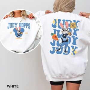 K&ouml;nnte beinhalten: Wei&szlig;es Sweatshirt mit einem Cartoon-Bild von Judy Hopps aus Zootopia. Die Vorderseite hat ein blaues Design mit dem Text "JUDY HOPPS" und einem Cartoon-Bild von Judy. Die R&uuml;ckseite hat eine gr&ouml;&szlig;ere Judy Hopps Grafik mit dem Text "JUDY" wiederholt.