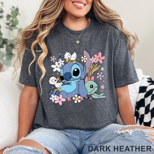K&ouml;nnte beinhalten: Dunkelgraues T-Shirt mit einem Cartoon-Stitch-Design, Blumen und anderen Figuren. Das T-Shirt hat einen Rundhalsausschnitt und kurze &Auml;rmel. Der Text "DARK HEATHER" befindet sich am unteren Rand des T-Shirts.