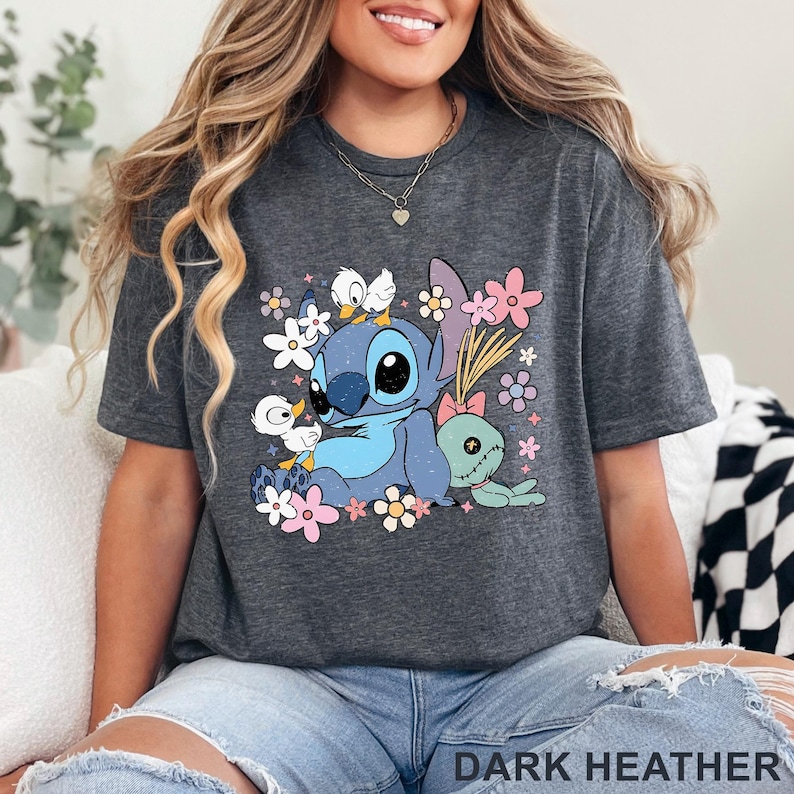 K&ouml;nnte beinhalten: Dunkelgraues T-Shirt mit einem Cartoon-Stitch-Design, Blumen und anderen Figuren. Das T-Shirt hat einen Rundhalsausschnitt und kurze &Auml;rmel. Der Text "DARK HEATHER" befindet sich am unteren Rand des T-Shirts.