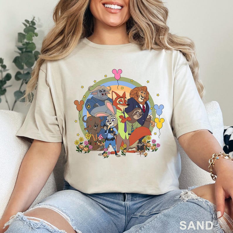 May include: Sand-colored t-shirt featuring a colorful graphic of Zootopia characters. The design includes Judy Hopps, Nick Wilde, and other characters in a rainbow-themed circle with flowers and Mickey Mouse-shaped balloons. The word "SAND" is printed at the bottom.