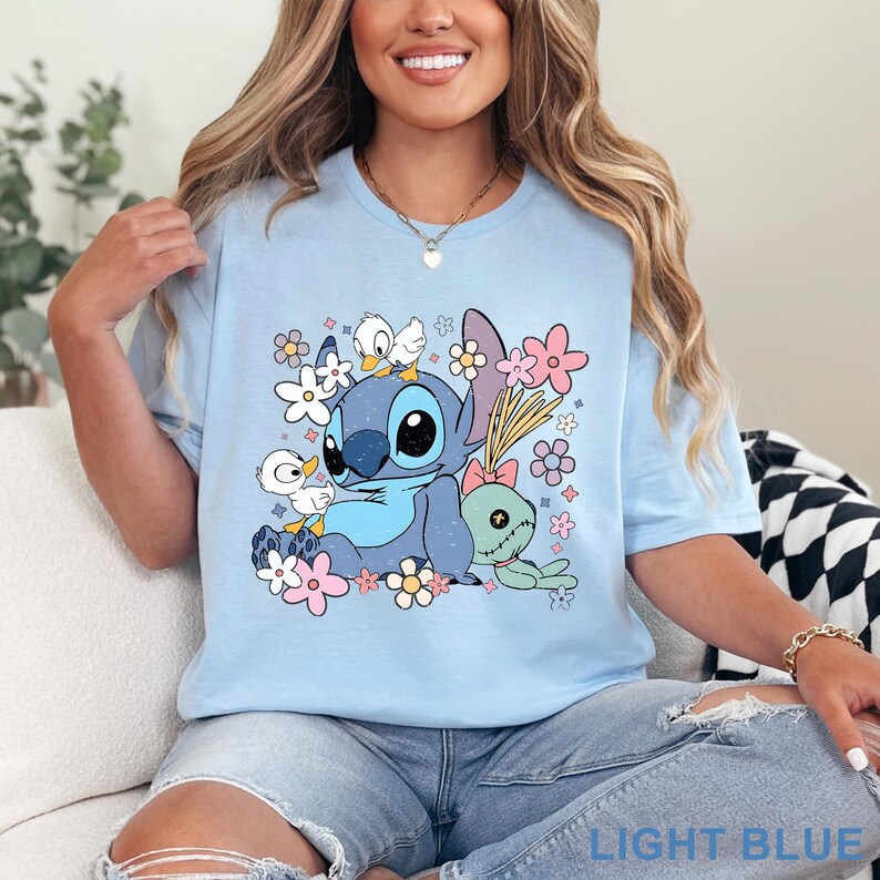 K&ouml;nnte beinhalten: Hellblaues T-Shirt mit einer Cartoonfigur, umgeben von Blumen und Enten. Der Text "LIGHT BLUE" befindet sich unten. Das Design umfasst rosa, wei&szlig;e und gelbe Blumen sowie eine gr&uuml;ne Schildkr&ouml;te.