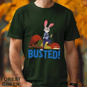 May include: Forest green t-shirt with a cartoon image of a rabbit police officer and a fox, with the word "BUSTED!" in blue. The rabbit is holding a small object. The shirt is a solid colour.
