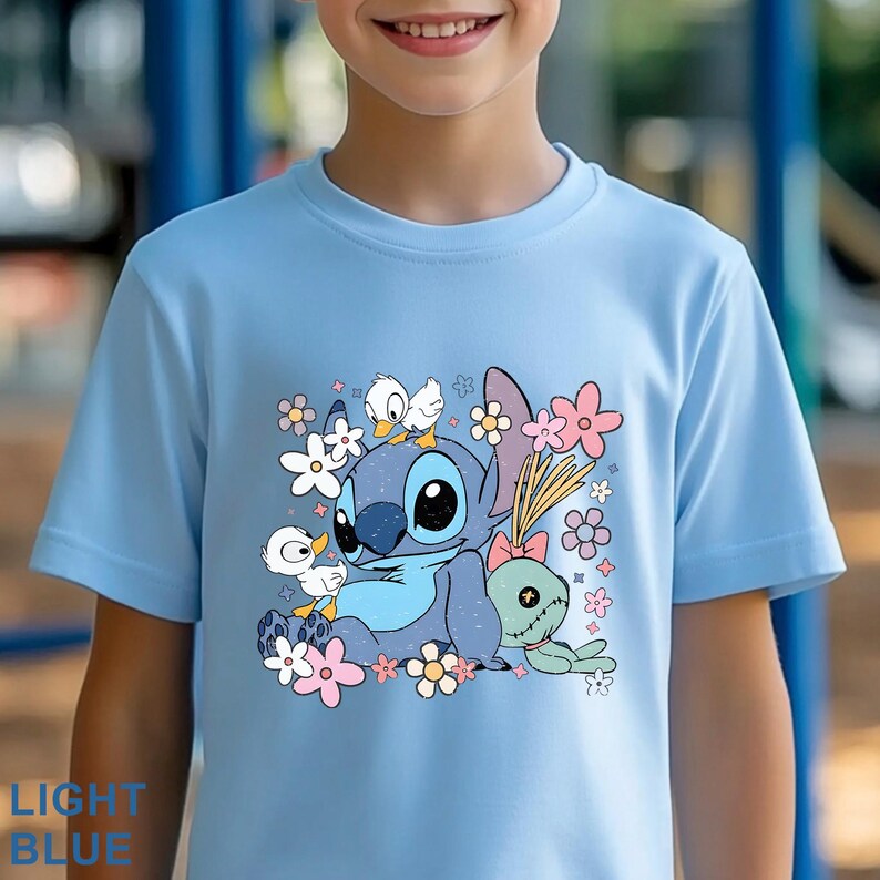 K&ouml;nnte beinhalten: Hellblaues T-Shirt mit einer Cartoonfigur, umgeben von Blumen und kleinen Entenk&uuml;ken. Das T-Shirt hat einen Rundhalsausschnitt und kurze &Auml;rmel. Der Text "LIGHT BLUE" ist unten links auf dem T-Shirt zu sehen.