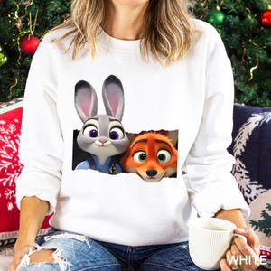 Może przedstawiać: Biała bluza z nadrukiem Judy Hopps i Nicka Wilde z Zwierzogrodu. Judy to szary kr&oacute;lik z fioletowymi oczami, a Nick to pomarańczowy lis z zielonymi oczami. Postacie wyłaniają się z efektu podartego papieru.
