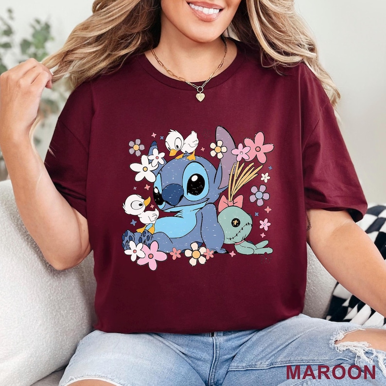 K&ouml;nnte beinhalten: Burgunderfarbenes T-Shirt mit einer Cartoonfigur in Blau, gro&szlig;en schwarzen Augen und zwei wei&szlig;en Entenk&uuml;ken. Das Design umfasst bunte Blumen und eine gr&uuml;ne Puppe. Das Wort "MAROON" ist unten aufgedruckt.