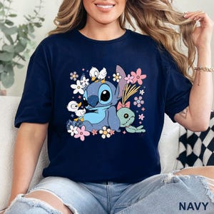 K&ouml;nnte beinhalten: Marineblaues T-Shirt mit einer Cartoon-Illustration von Stitch, Blumen und Enten. Die Grafik enth&auml;lt eine gr&uuml;ne Puppe und das Wort "NAVY". Das Design verwendet Blaut&ouml;ne, Rosa und Wei&szlig;.