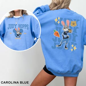 K&ouml;nnte beinhalten: Carolina-blaues Sweatshirt mit Judy Hopps aus Zootopia. Auf der Vorderseite befindet sich eine Grafik von Judy Hopps im Mickey-Mouse-Stil mit dem Text "Judy Hopps". Auf der R&uuml;ckseite befindet sich ein gr&ouml;&szlig;eres Bild von Judy Hopps in ihrer Polizeiuniform mit dem Text "Judy Joy".