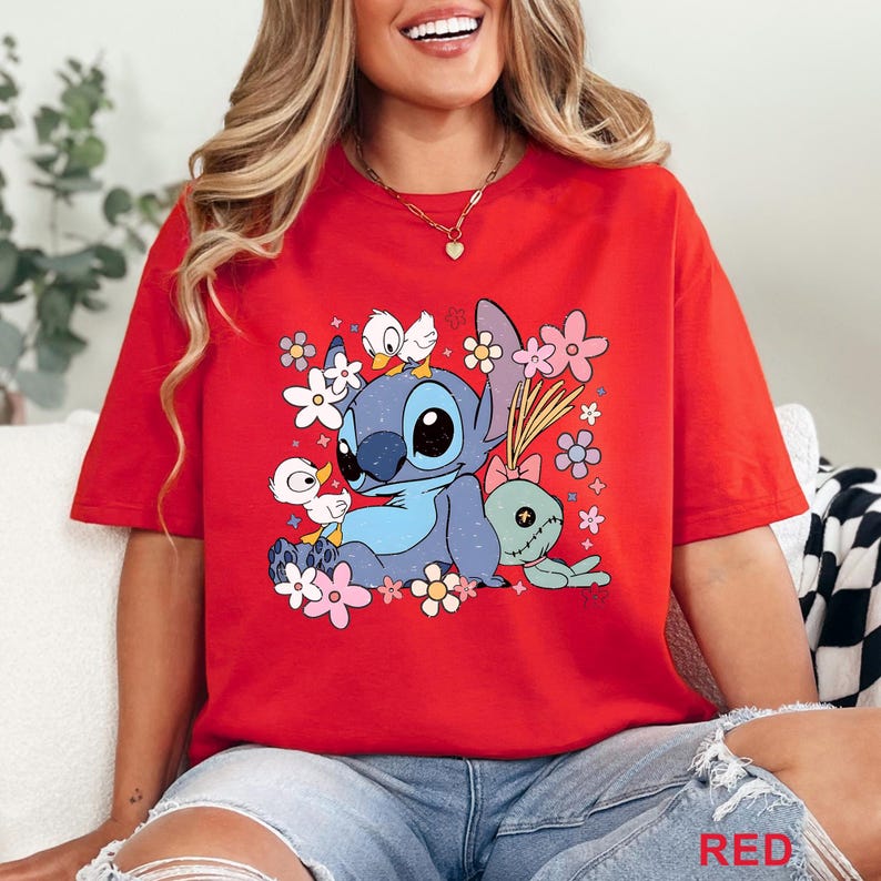 K&ouml;nnte beinhalten: Rotes T-Shirt mit einer blauen Cartoonfigur mit gro&szlig;en Augen, umgeben von Blumen und kleinen Entenk&uuml;ken. Die Grafik enth&auml;lt eine gr&uuml;ne Puppe und das Wort "RED" unten.
