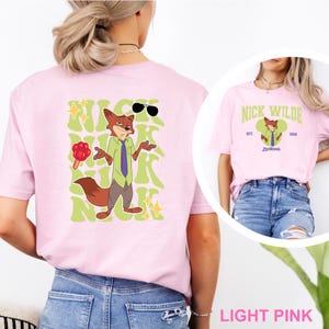 May include: Light pink t-shirt featuring a cartoon fox character with sunglasses, a green shirt, and a purple tie. The text "NICK WILDE" is printed in green, along with the word "Zootopia". The shirt is a light pink color.