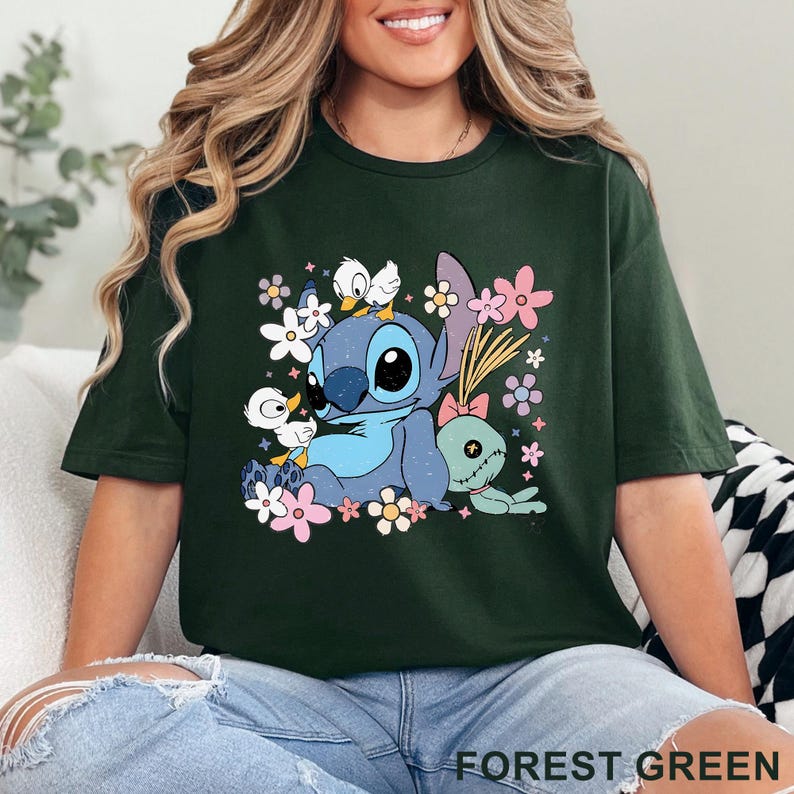 K&ouml;nnte beinhalten: Forstgr&uuml;nes T-Shirt mit einer blauen Cartoonfigur, gro&szlig;en Augen, Blumen und Entenk&uuml;ken. Eine gr&uuml;ne Puppe mit einer rosa Schleife ist ebenfalls vorhanden. Der Text "FOREST GREEN" befindet sich unten.
