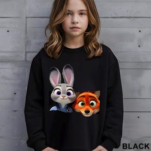 Może przedstawiać: Czarna bluza z ilustracją Judy Hopps, szarego kr&oacute;lika, i Nicka Wilde, pomarańczowego lisa, z filmu Zwierzogr&oacute;d. Słowo "BLACK" jest wydrukowane na dole po prawej stronie na biało. Postacie mają szczeg&oacute;łowe cechy kresk&oacute;wkowe.