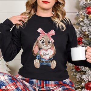 May include: Black sweatshirt with a cartoon bunny holding a heart that says "love". The bunny has a pink bow and is wearing a blue uniform. The word "BLACK" is printed at the bottom of the image.