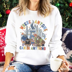 May include: White sweatshirt featuring a colorful graphic of a city skyline and cartoon characters. The text "WHERE ANYONE CAN BE ANYTHING" is displayed above the characters. The sweatshirt is a crew neck style.