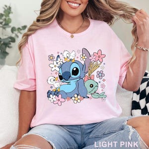 K&ouml;nnte beinhalten: Hellrosa T-Shirt mit einer Cartoonfigur mit blauem Fell, gro&szlig;en schwarzen Augen und einem hellblauen Bauch, umgeben von Blumen und kleinen wei&szlig;en Enten. Der Text "LIGHT PINK" befindet sich unten.