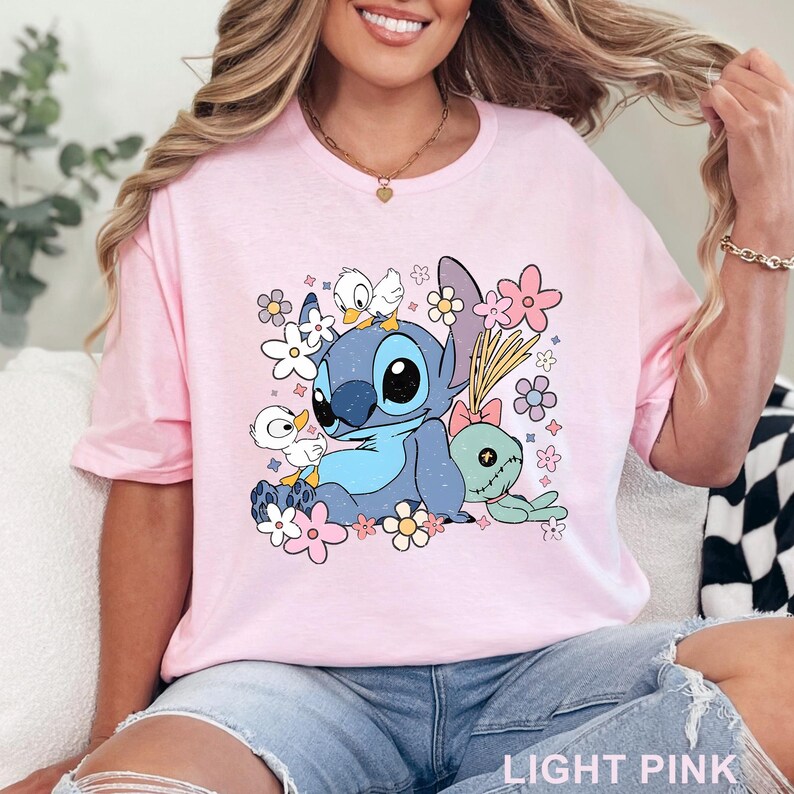 K&ouml;nnte beinhalten: Hellrosa T-Shirt mit einer Cartoonfigur mit blauem Fell, gro&szlig;en schwarzen Augen und einem hellblauen Bauch, umgeben von Blumen und kleinen wei&szlig;en Enten. Der Text "LIGHT PINK" befindet sich unten.