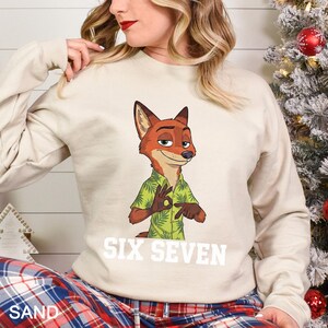 May include: A sand-colored sweatshirt featuring a cartoon fox character in a green Hawaiian shirt, making an 'okay' hand gesture. The text "SIX SEVEN" is printed in white below the character. The sweatshirt is part of a casual clothing collection.