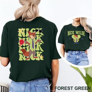 May include: Forest green t-shirt featuring a cartoon fox character with sunglasses and the text "Nick Wilde" in a stylized font. The design includes a lollipop and the character's name. The shirt is a casual wear item.