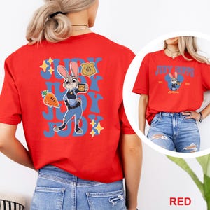 May include: Red t-shirt featuring a cartoon image of Judy Hopps from Zootopia. The design includes Judy in a police uniform, the words "Judy Hopps" in blue, and other Zootopia-themed graphics. The shirt is short-sleeved.