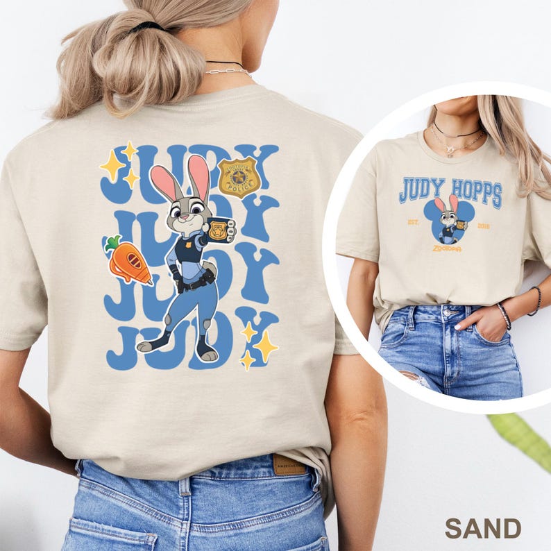 May include: Sand-colored t-shirt featuring a cartoon image of Judy Hopps from Zootopia. The back of the shirt has a blue design with Judy Hopps, a carrot, and a police badge. The front of the shirt has the text "Judy Hopps" and a cartoon image of Judy.