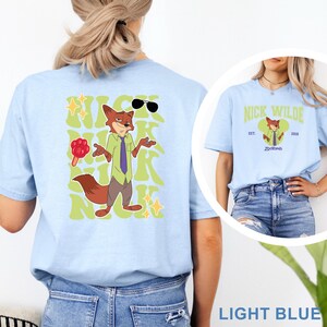 May include: Light blue t-shirt featuring a cartoon fox character with sunglasses and the text "NICK" in a retro font. The shirt also has a smaller image of the character and the text "NICK WILDE" and "Zootopia".