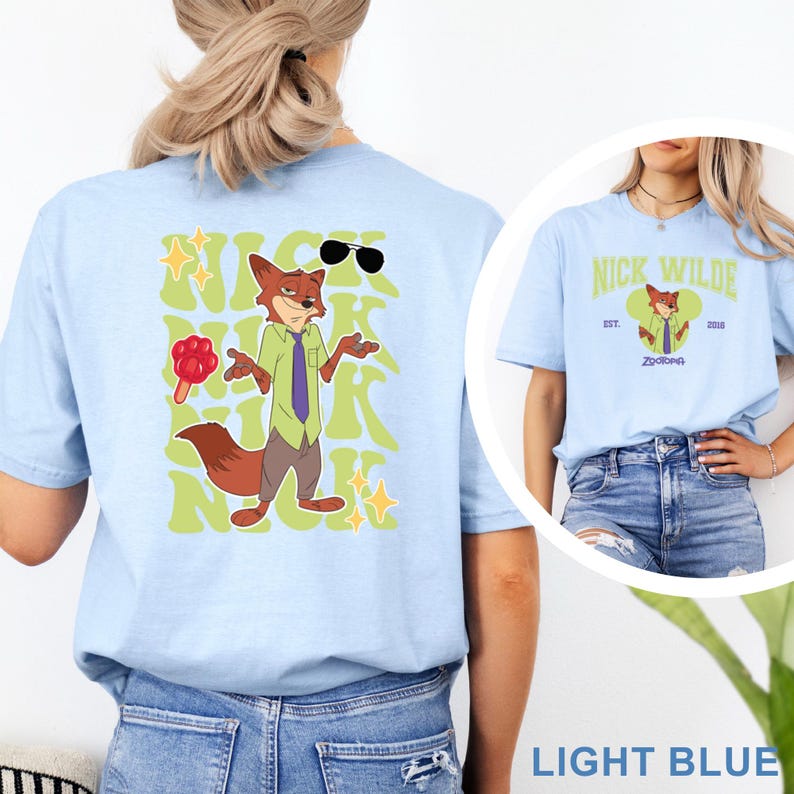 May include: Light blue t-shirt featuring a cartoon fox character with sunglasses and the text "NICK" in a retro font. The shirt also has a smaller image of the character and the text "NICK WILDE" and "Zootopia".